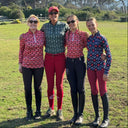 Four people in equestrian attire standing on a grassy field with mountains in the background